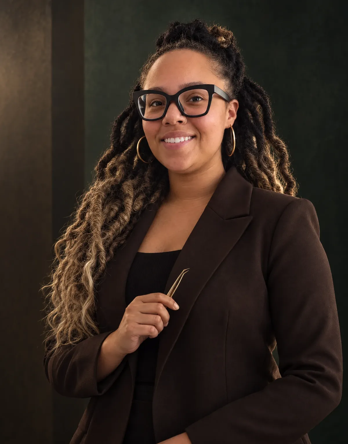 Desiree, the lash and brow artist at Central Oasis, smiling in a dark brown blazer and holding lash tweezers