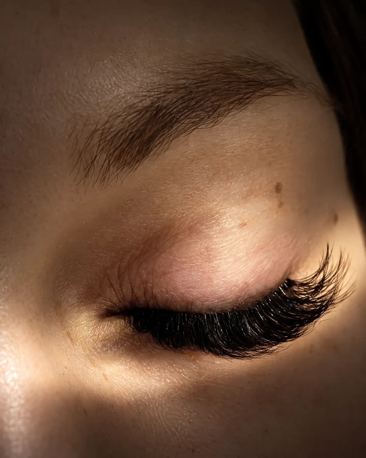 A finished set of dark lash extensions on closed eyes, lit by a soft band of warm light
