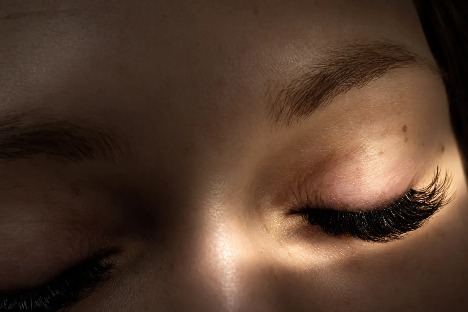 A finished set of dark lash extensions on closed eyes, lit by a soft band of warm light