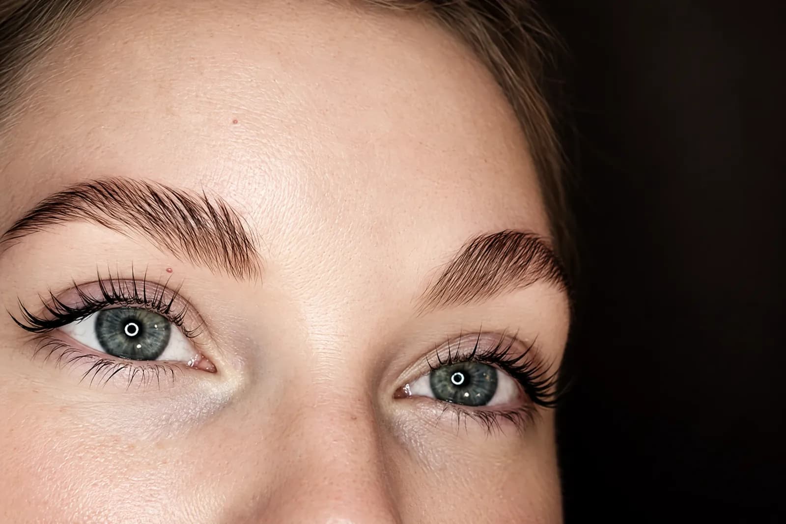 Brushed-up, feathered brows above green eyes and a clean, dark lash line, close up against a dark background
