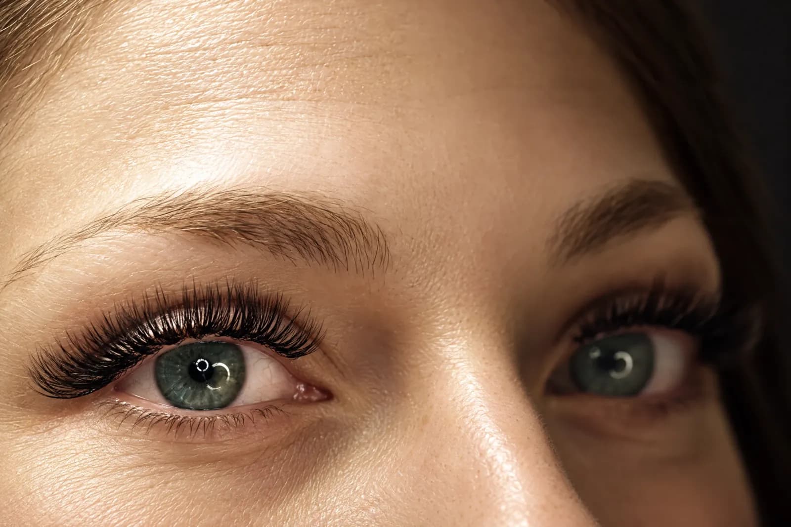 Full, dark lash extensions on green eyes glancing up, with soft brows above, in warm light
