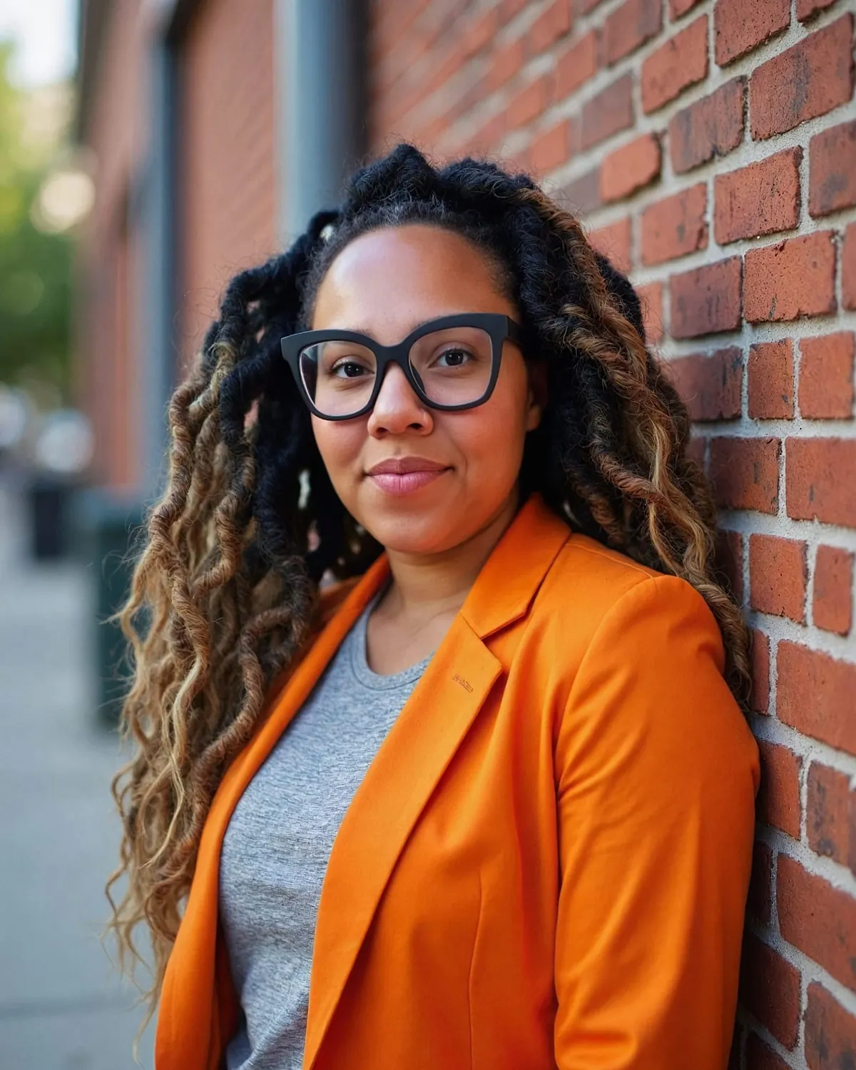 Desiree, the lash and brow artist at Central Oasis, smiling in dark-framed glasses and an orange blazer over a grey tee, leaning against a red brick wall